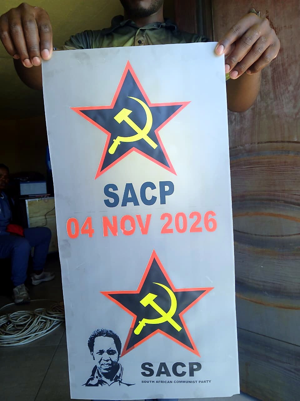 F-DTF/A3 DTF print: A3 transfer film sheet with SACP star, hammer-and-sickle and '04 NOV 2026' text held up in workshop, ready for H-PRESS/6080D pressing (staff still)