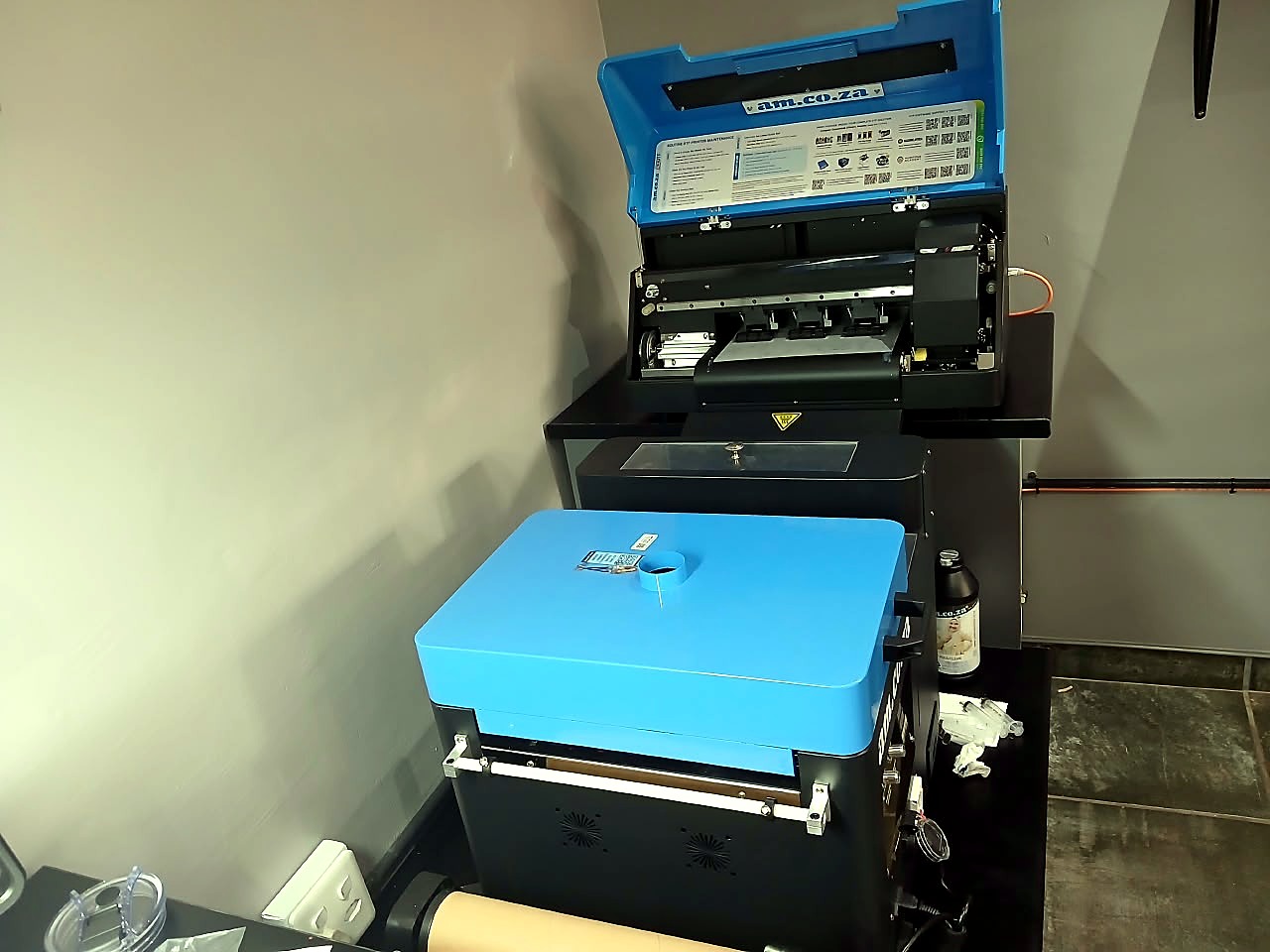 F-DTF/A3 DTF: am.co.za A3 DTF printer with blue lid open (XP600 carriage, rails and quick-guide sticker visible) on a stand behind the blue H-DTF/POWDER/300 rolled-film powder shaking and baking unit (staff still)