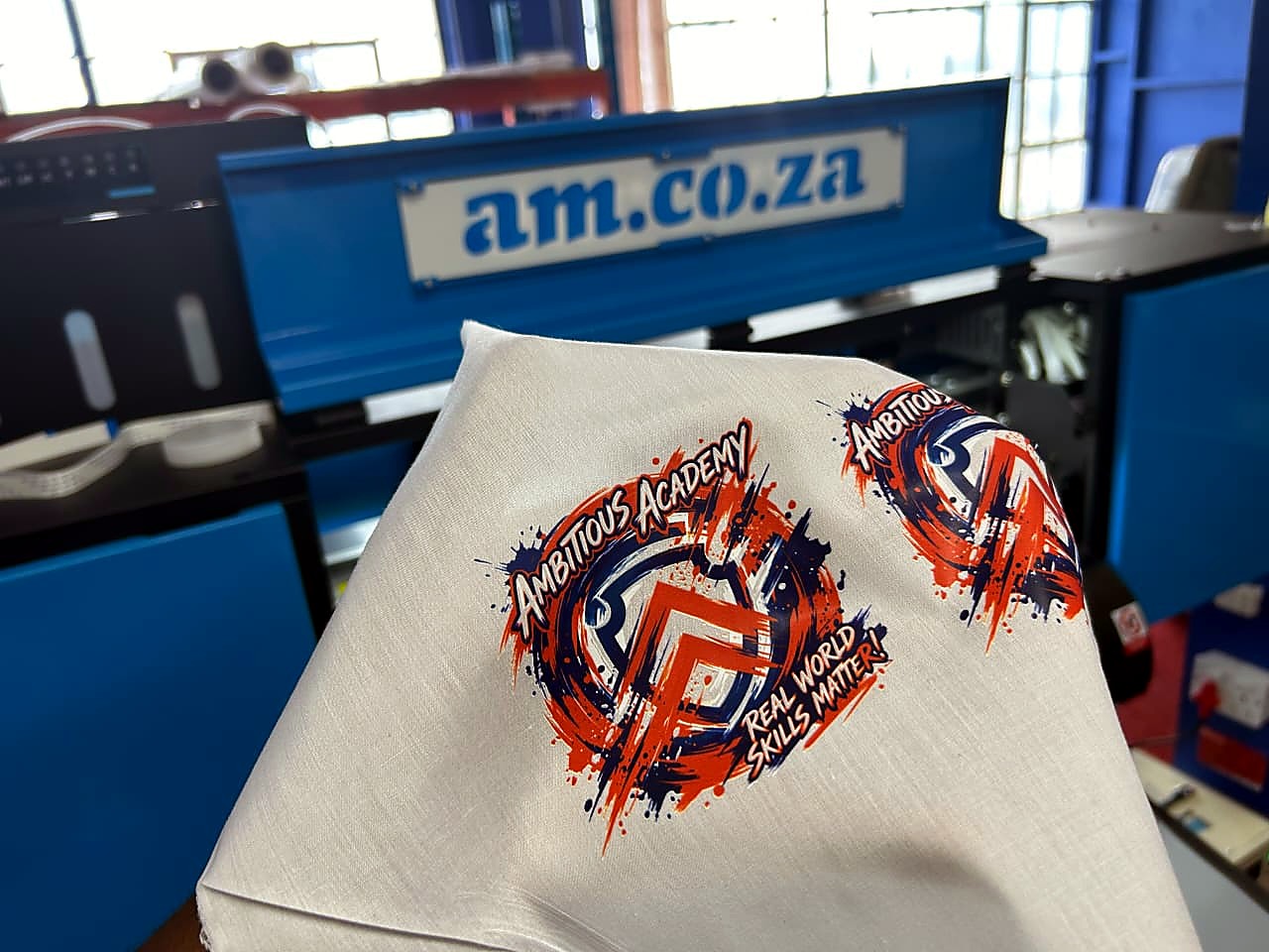 H-DTF/600 DTF: full-colour 'Ambitious Academy – Real World Skills Matter!' logo transfers heat-pressed onto white fabric, held in front of the blue am.co.za DTF printer (staff still)