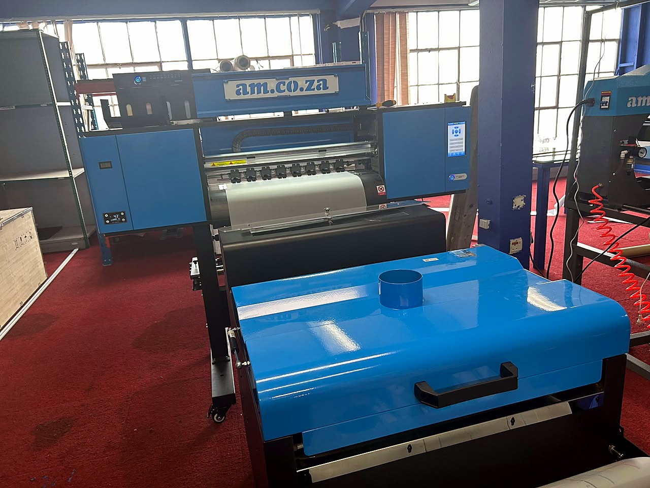 H-DTF/600 DTF: blue/black am.co.za 600mm DTF printer with touch panel, white DTF film on the take-up and the blue powder shaker/curing unit in front, in the AM.CO.ZA showroom (staff still)