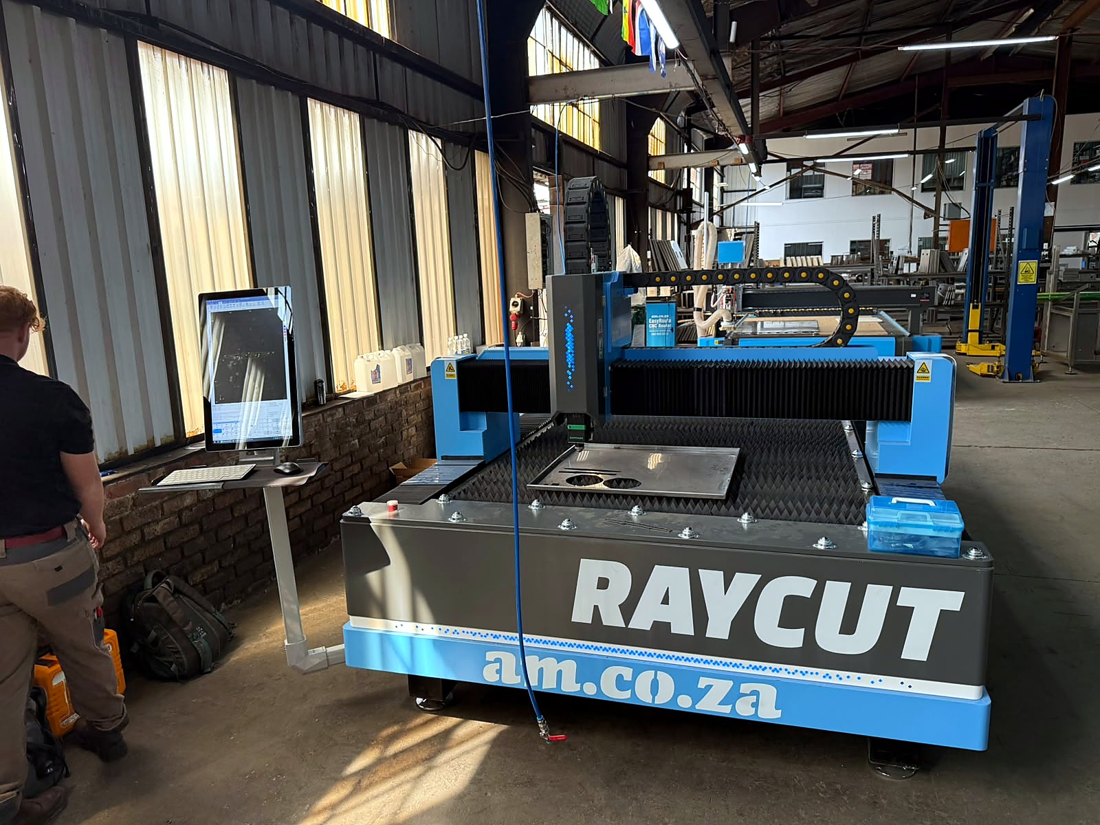 LF-1530/3000M RayCUT fiber laser: blue/black RAYCUT am.co.za machine in workshop with cut steel plate on slat bed, operator at standing control PC (staff still)