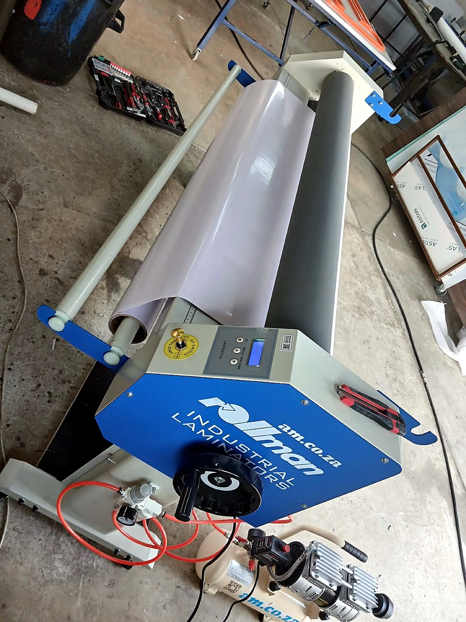 RM-1520 Rollman laminator: blue 'Rollman Industrial Laminators am.co.za' side panel with handwheel, LCD controls, red air lines and air compressor beside it (staff still)