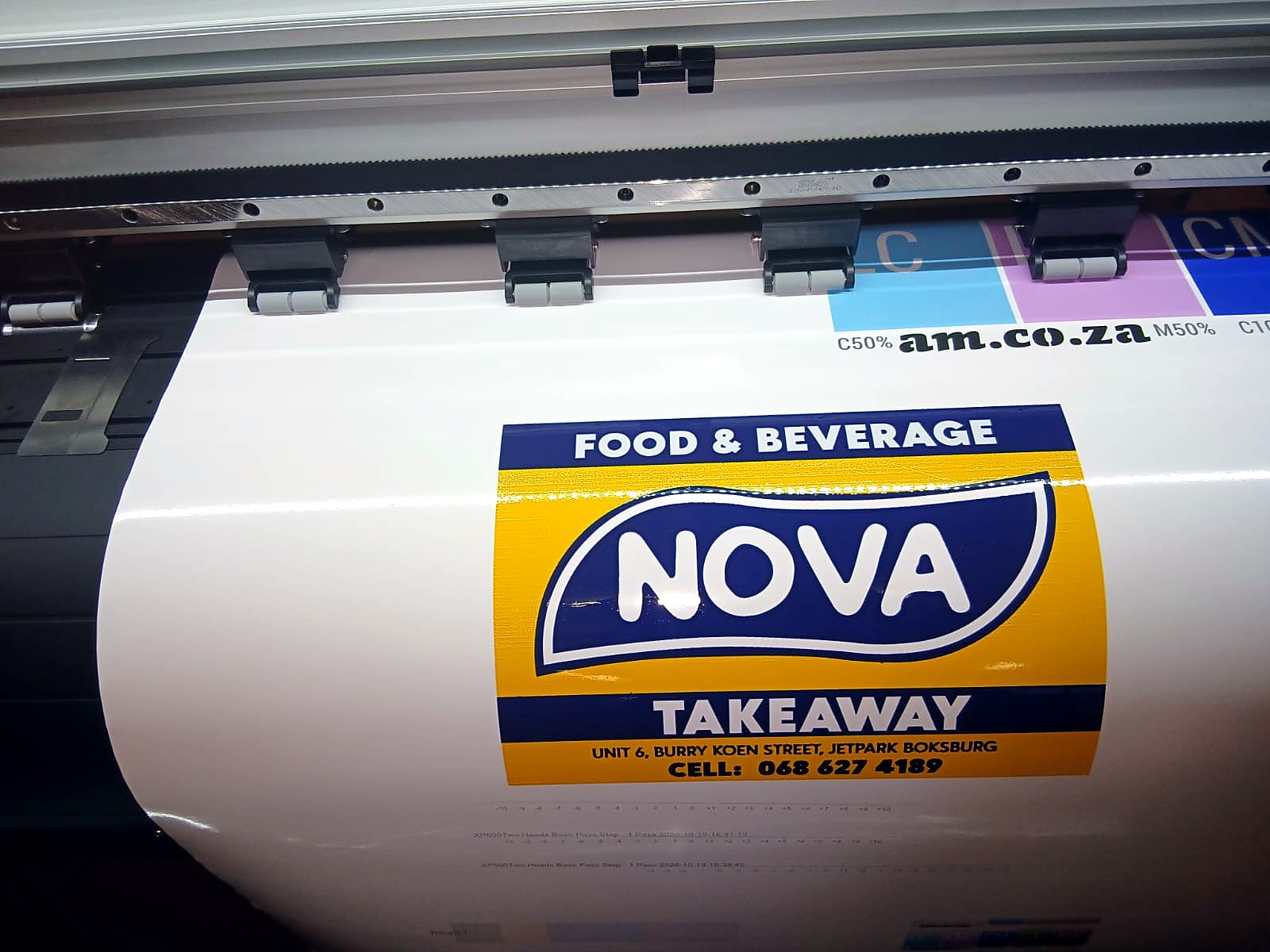 F1-1800 FastCOLOUR: full-colour takeaway shop signage print on gloss white vinyl coming off the platen under the pinch rollers, with am.co.za C/M/CM colour-bar strip above (staff still)