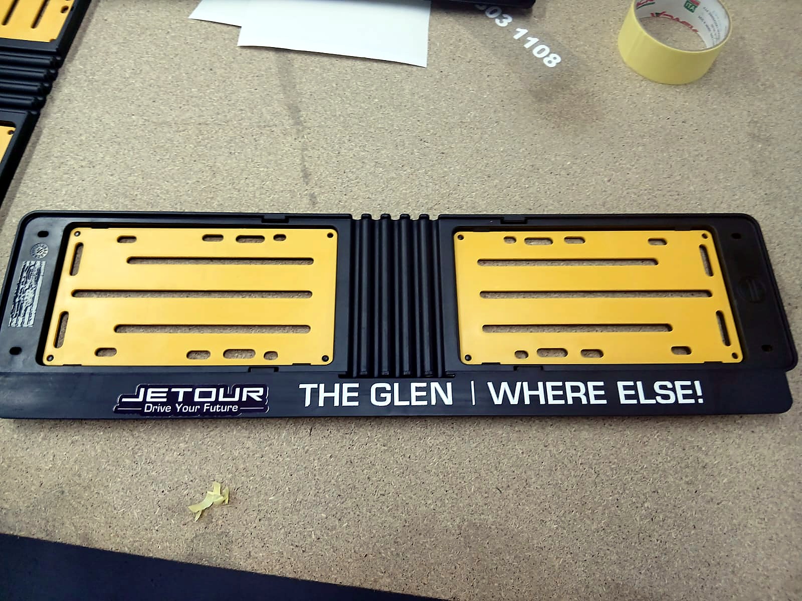F-UVDTF/600 UV DTF result: black number-plate holder with applied 'JETOUR Drive Your Future | THE GLEN | WHERE ELSE!' transfer on the bottom strip (staff still)