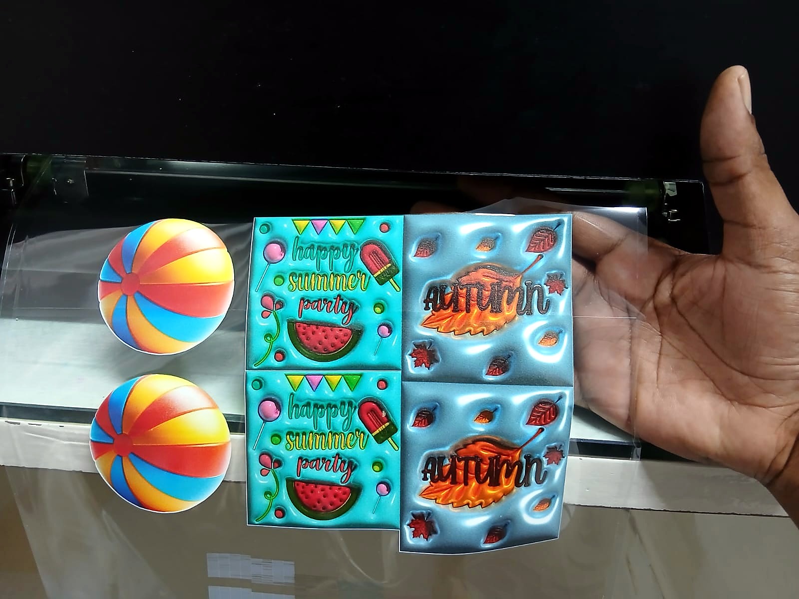 F-UVDTF/A3 UV-DTF: laminated transfer film held up to the light showing glossy 3D-effect UV-DTF stickers – beach balls, 'happy summer party' and 'AUTUMN' leaf designs (staff still)
